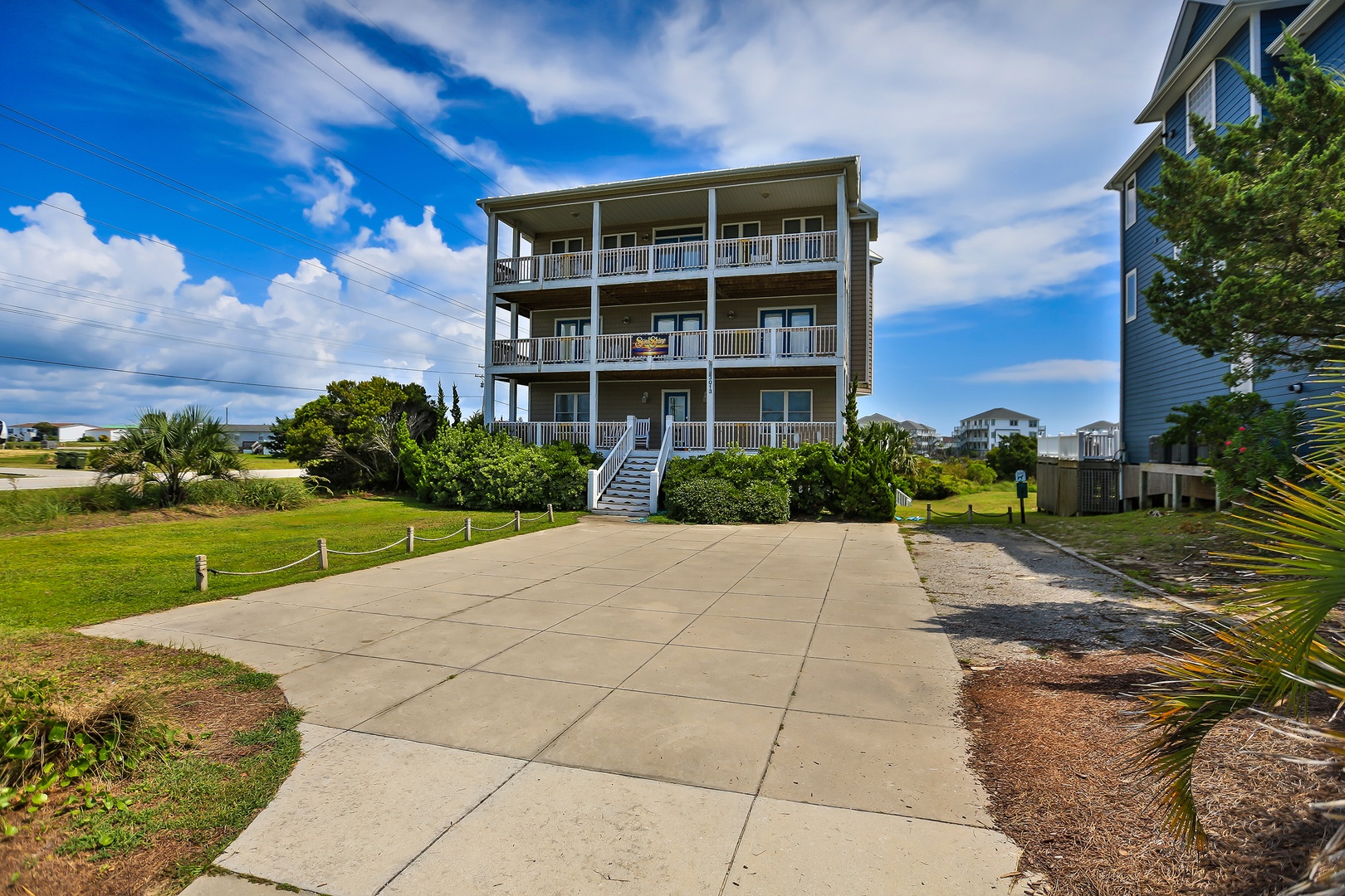 Three-story coastal rental featuring wraparound balconies and tropical landscaping in a peaceful neighborhood setting.