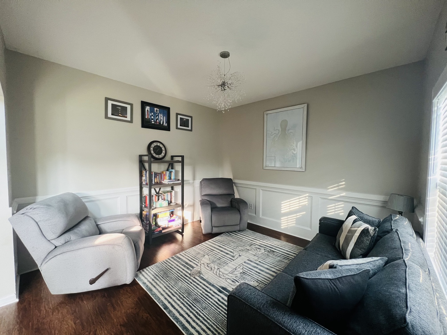Sink into plush seating in your elegant living space, where natural light dances through blinds and a sparkling chandelier creates the perfect atmosphere for relaxation.