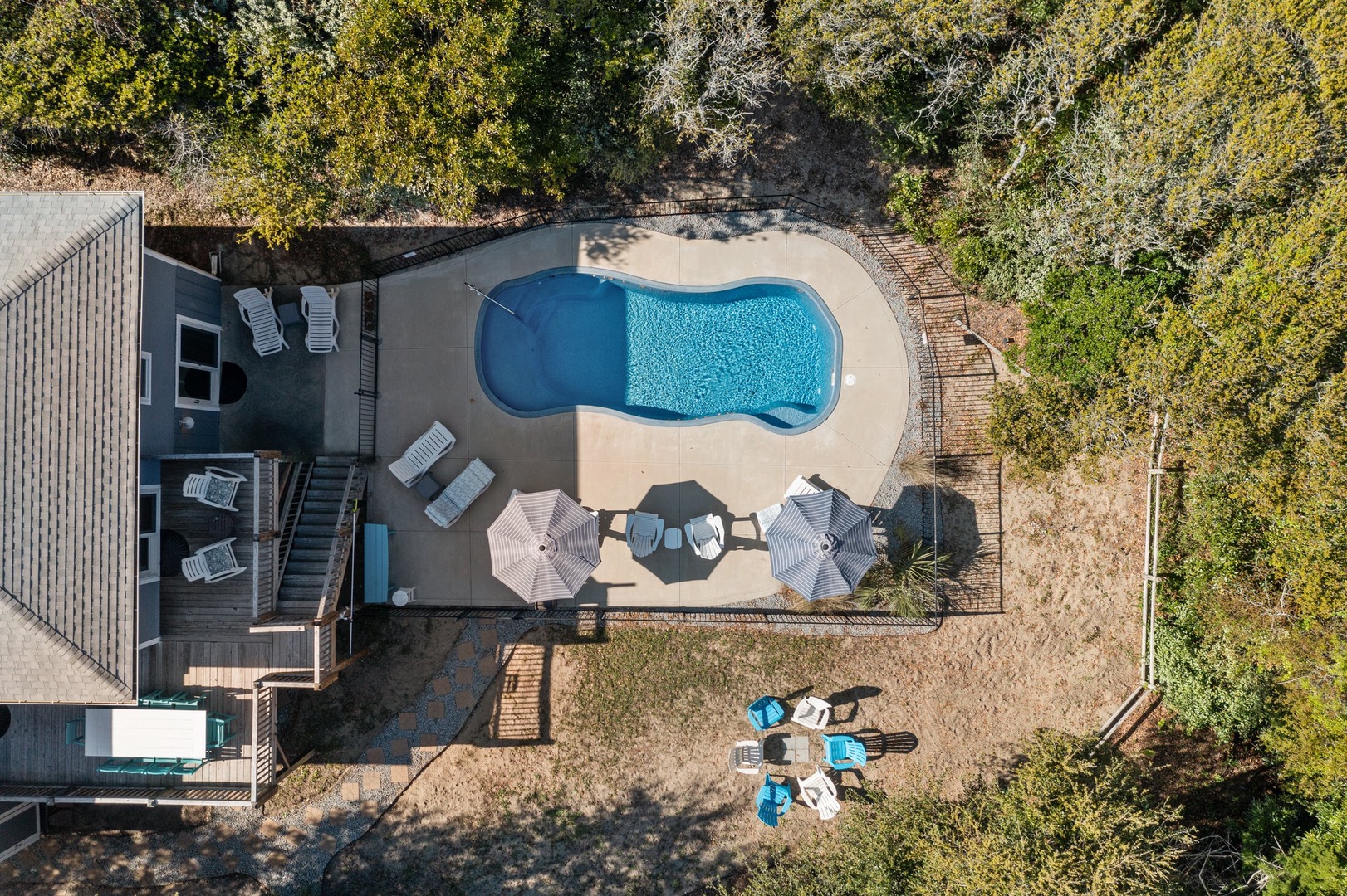 Aerial view of vacation rental with curved pool, lounge chairs, and umbrellas surrounded by lush wooded landscape.