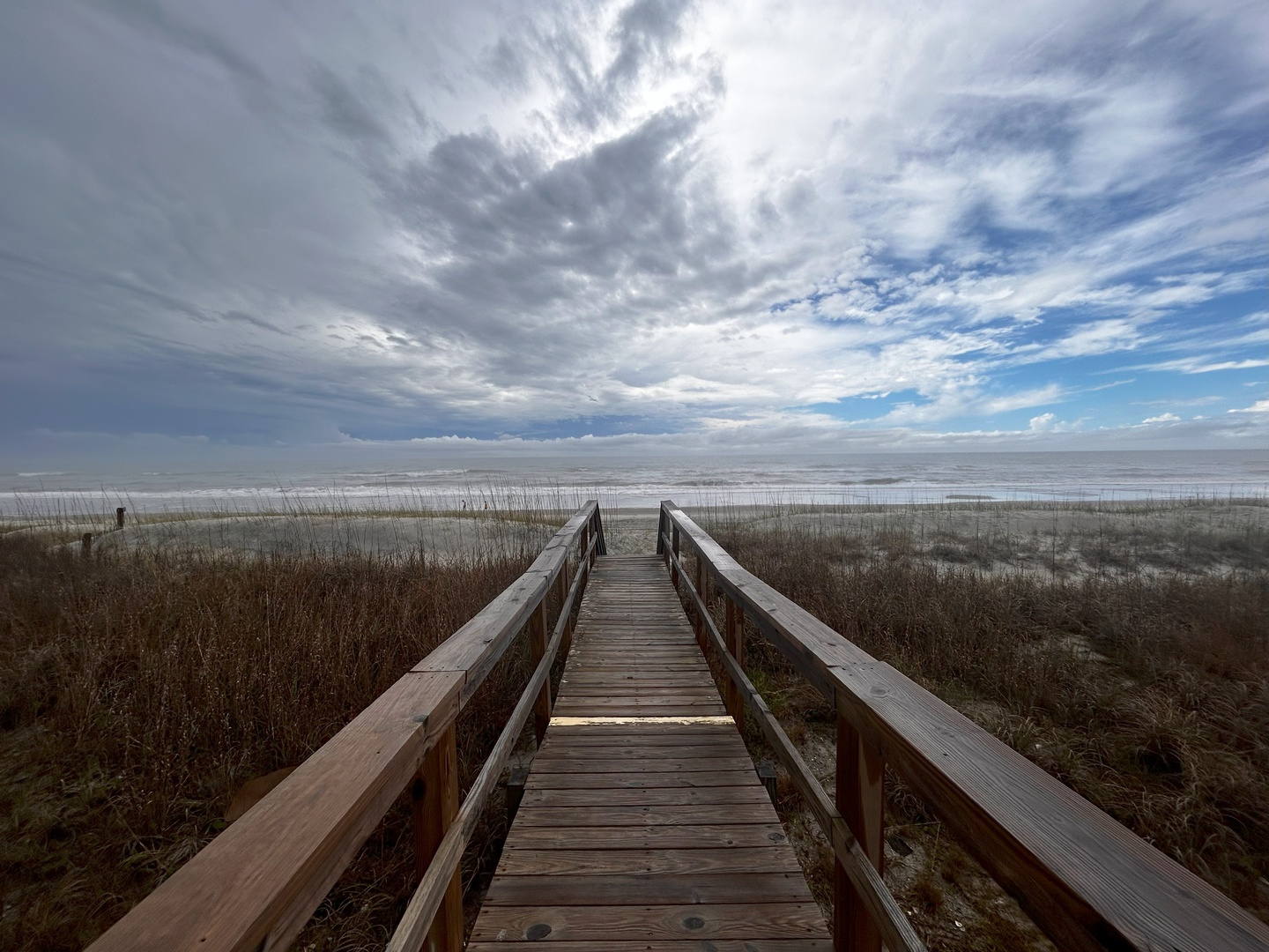 Wooden boardwalk leads through coastal dunes to pristine beach under dramatic cloudy skies.