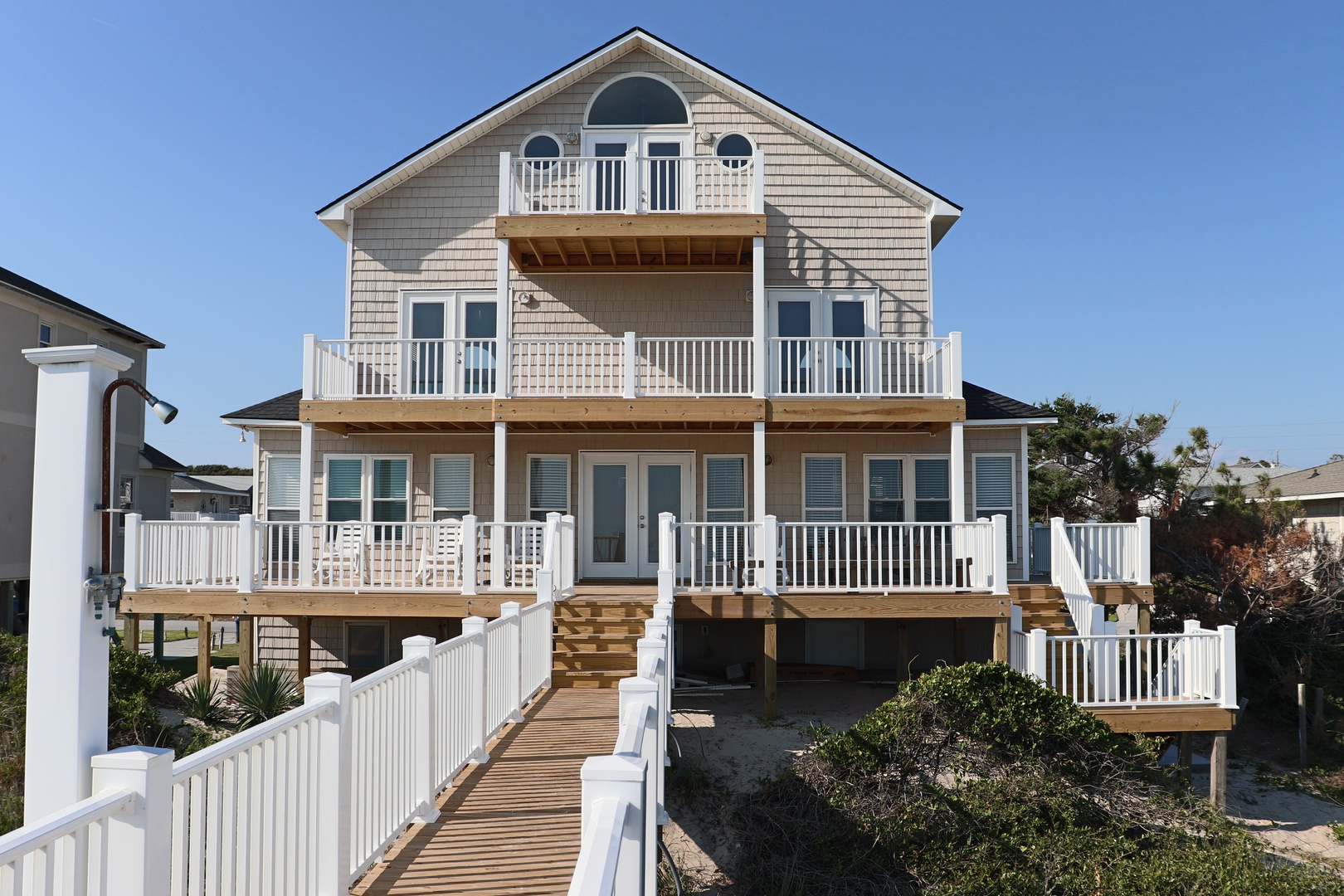 Multi-level coastal home with wraparound decks and balconies, surrounded by lush landscaping under clear blue skies.