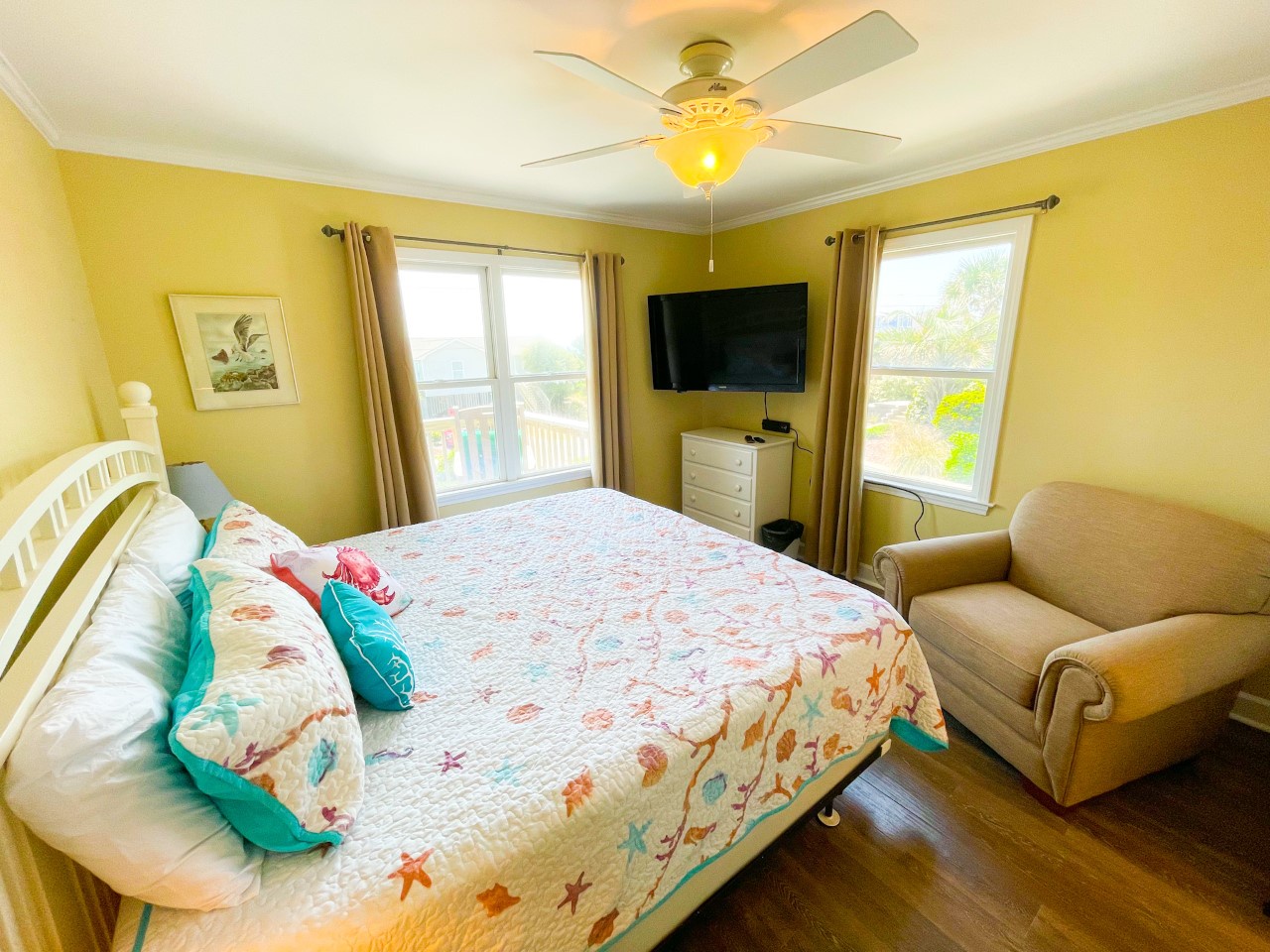 Your cheerful bedroom features a comfortable bed with coastal-themed bedding, cozy armchair, TV entertainment, and natural light streaming through windows with lovely outdoor views.