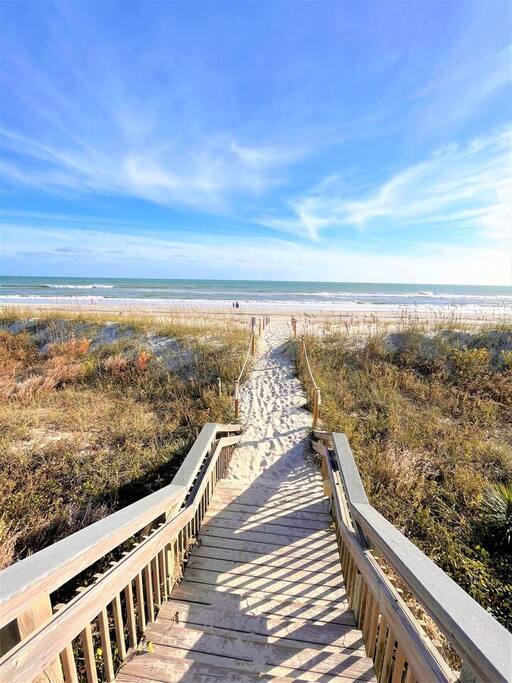 Wooden boardwalk leads through coastal dunes to pristine beach shoreline under expansive blue sky.