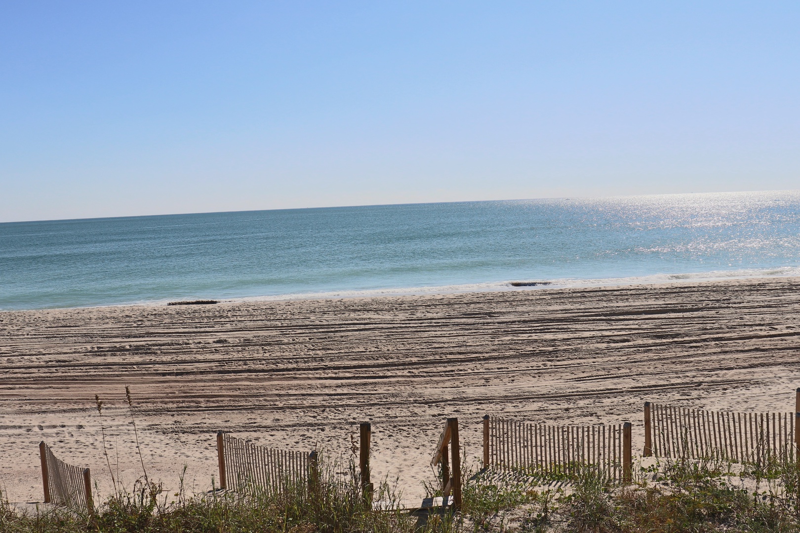 Pristine sandy beach stretches along turquoise waters under clear blue skies, with protective dune fencing maintaining this coastal paradise.
