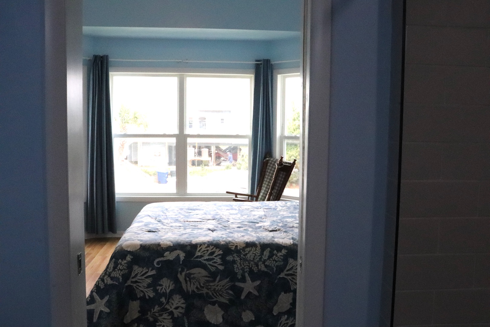 Your serene bedroom features soft blue tones, a comfortable bed with coastal-themed linens, and bright windows framing peaceful neighborhood views.