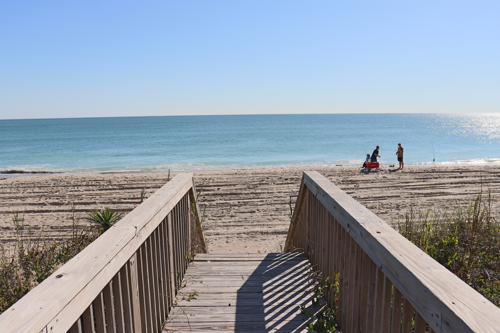 Pristine beach with wooden boardwalk leading to crystal-clear waters and soft sand where families enjoy peaceful seaside moments.
