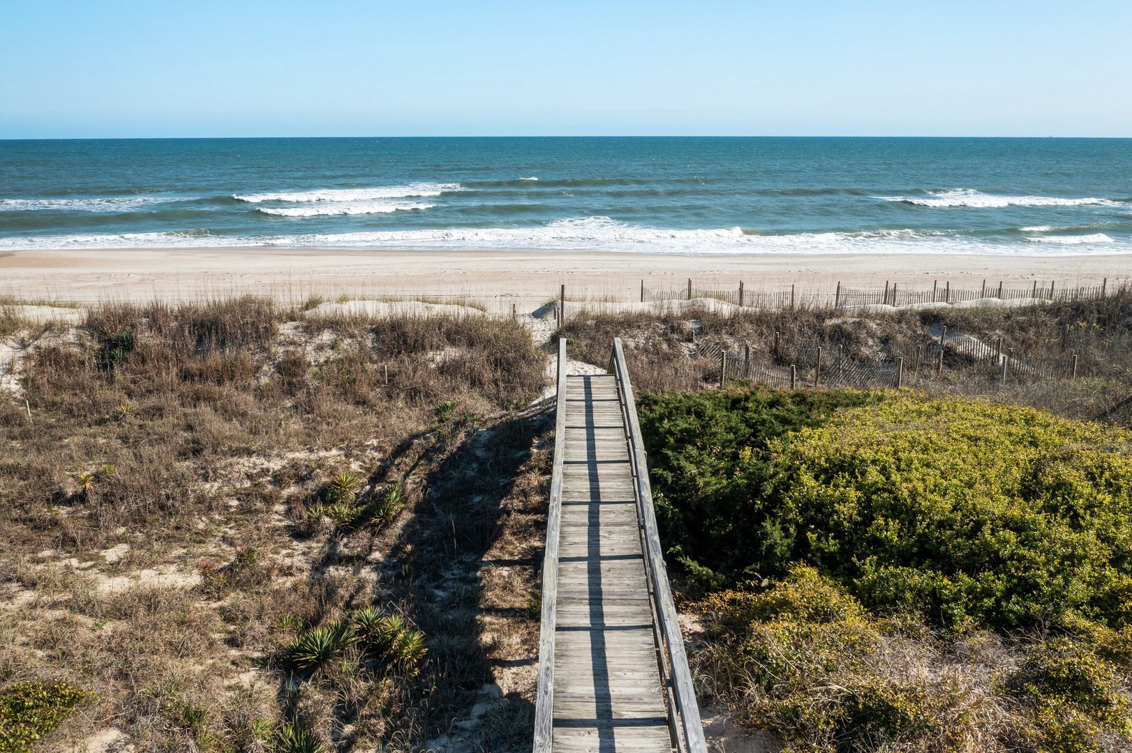 Wooden boardwalk leads across coastal dunes to pristine sandy beach with rolling ocean waves.