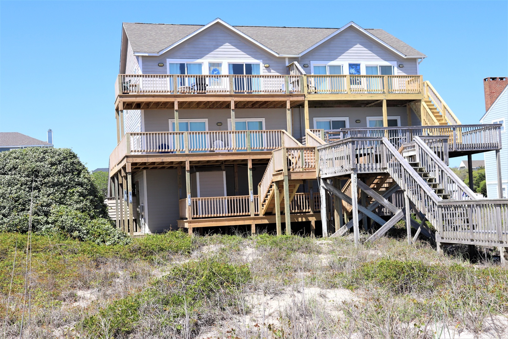 Multi-level beach house with expansive decks and wooden stairs, surrounded by coastal dunes and vegetation.