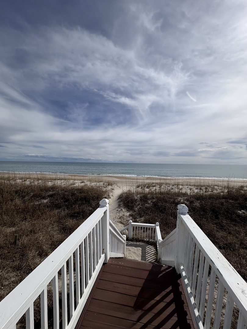 White boardwalk leads across coastal dunes to pristine sandy beach and ocean waves under dramatic cloudy skies.