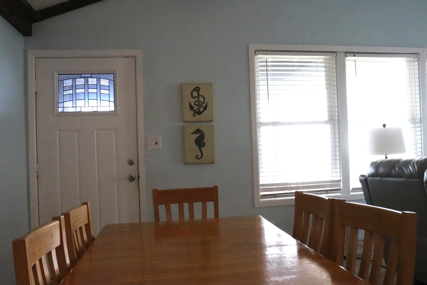 The dining table awaits your group meals and vacation memories, bathed in natural light from coastal-themed windows near the entrance.