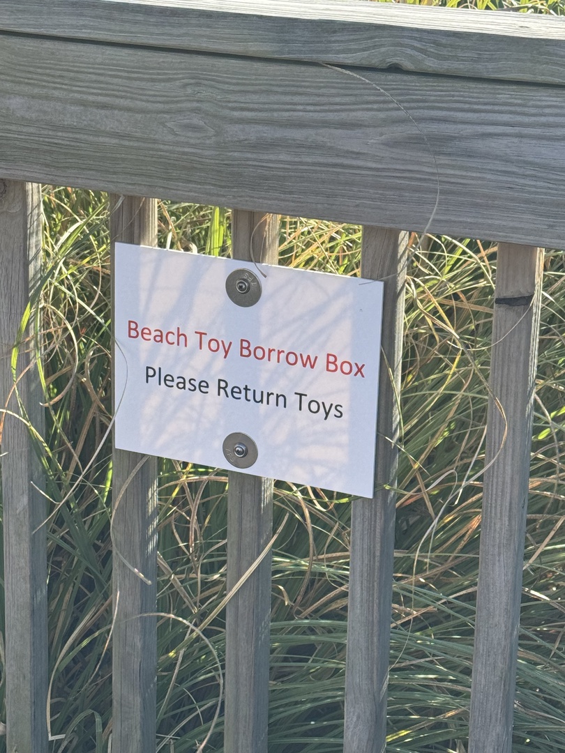 Beach Toy Borrow Box sign attached to wooden fence with natural grass backdrop.