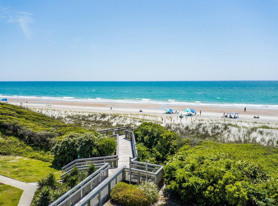 Pristine beach stretches along turquoise waters, with boardwalk access through coastal dunes and native vegetation.