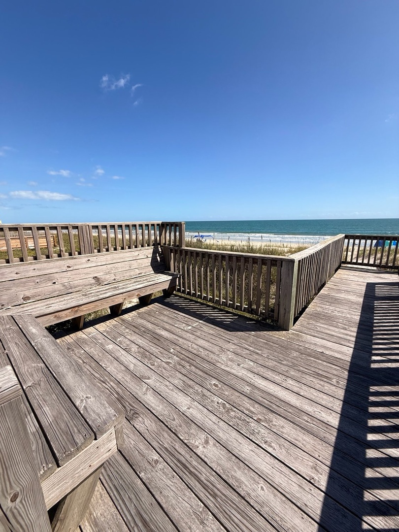 An oceanfront deck with built-in bench seating offering direct beach access. Weathered wood planks and railings frame views of dunes, rolling surf, and endless blue sky—an ideal spot to unwind with the sound of waves.