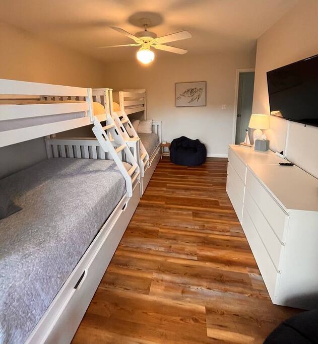 Your comfortable bunk room features white wooden beds, warm hardwood floors, and modern amenities including a flat-screen TV and ceiling fan for perfect relaxation.