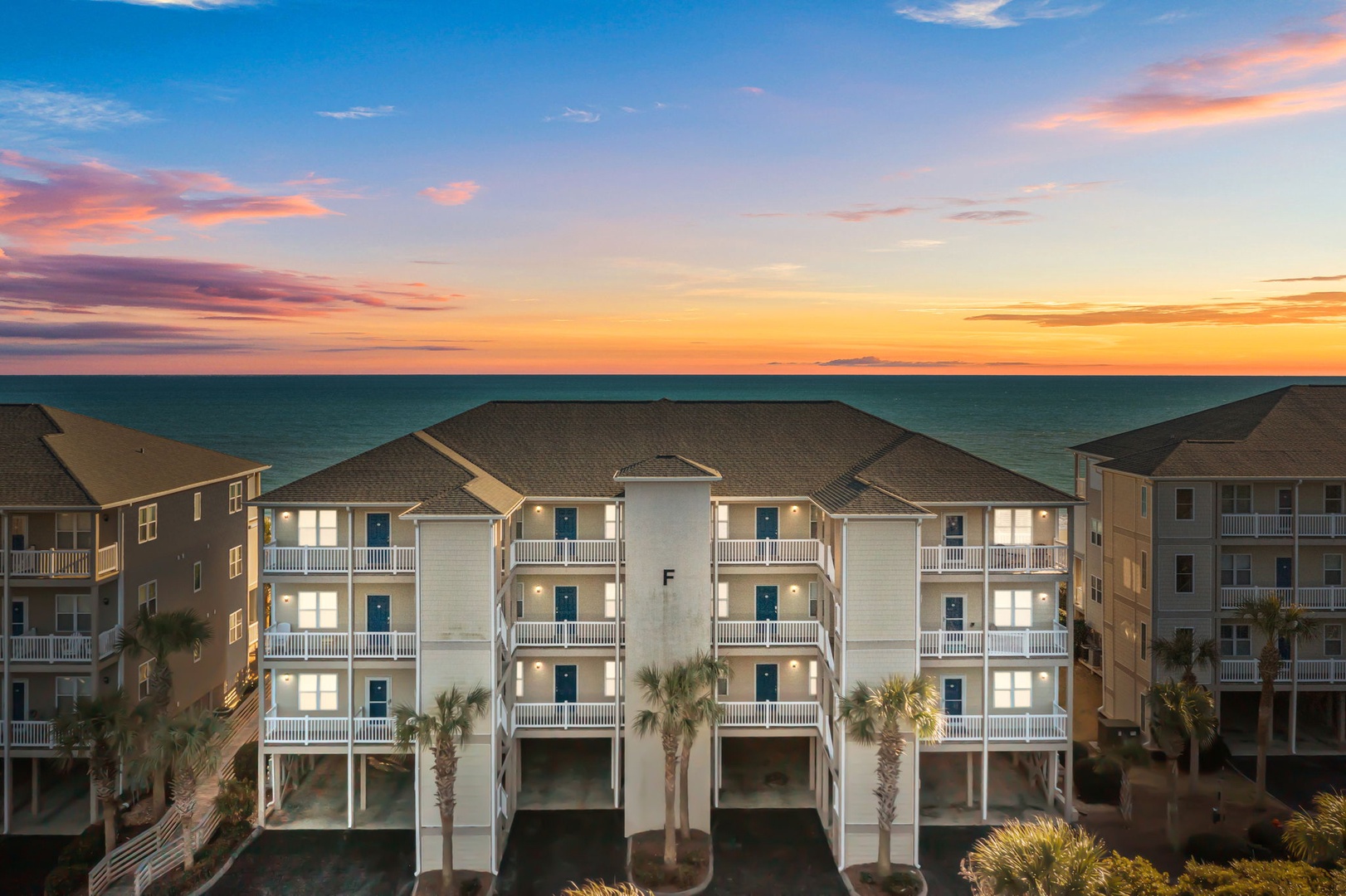 Oceanfront property complex featuring multiple buildings with balconies and palm trees during a stunning sunset.
