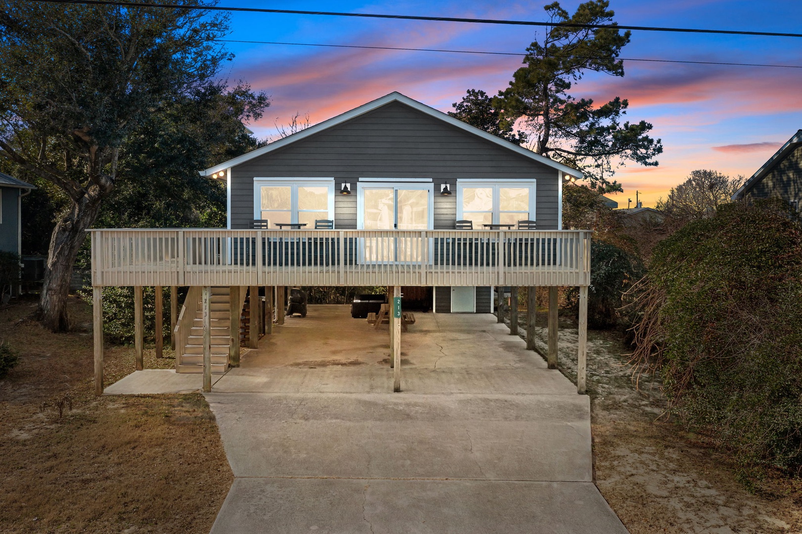 Beach cottage on stilts with wraparound deck and covered parking underneath, surrounded by coastal landscape during sunset.