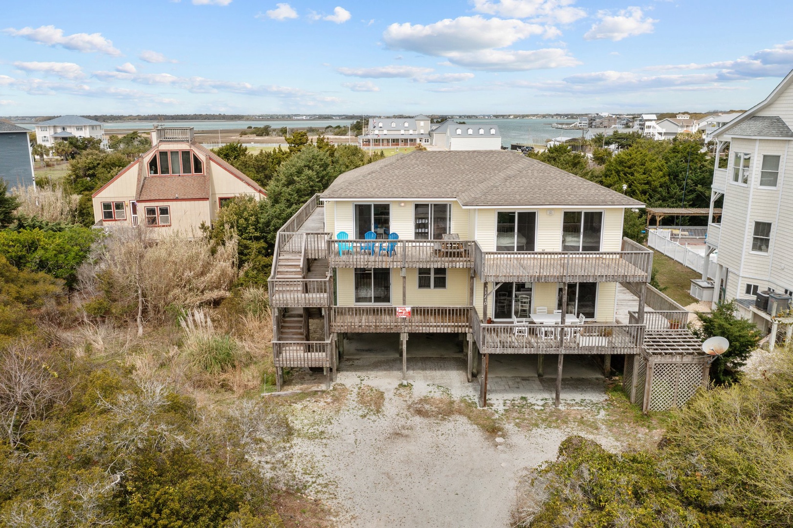 Elevated coastal home with expansive decks overlooks peaceful waterfront community, surrounded by lush vegetation and neighboring beach houses.