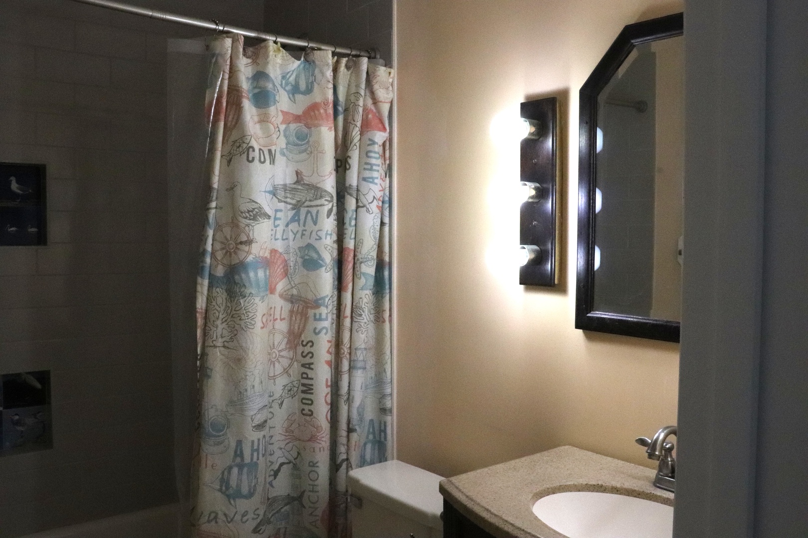 Your private bathroom features a vanity with granite countertop, shower-tub combo with nautical-themed curtain, and wall-mounted sconce lighting for warm ambiance.