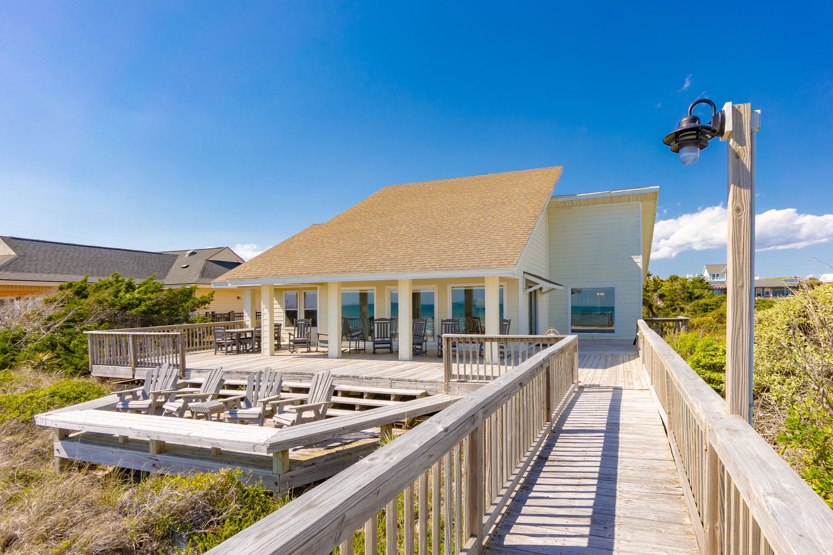 Charming beachfront cottage with expansive deck featuring lounge chairs and outdoor dining space, surrounded by coastal vegetation.