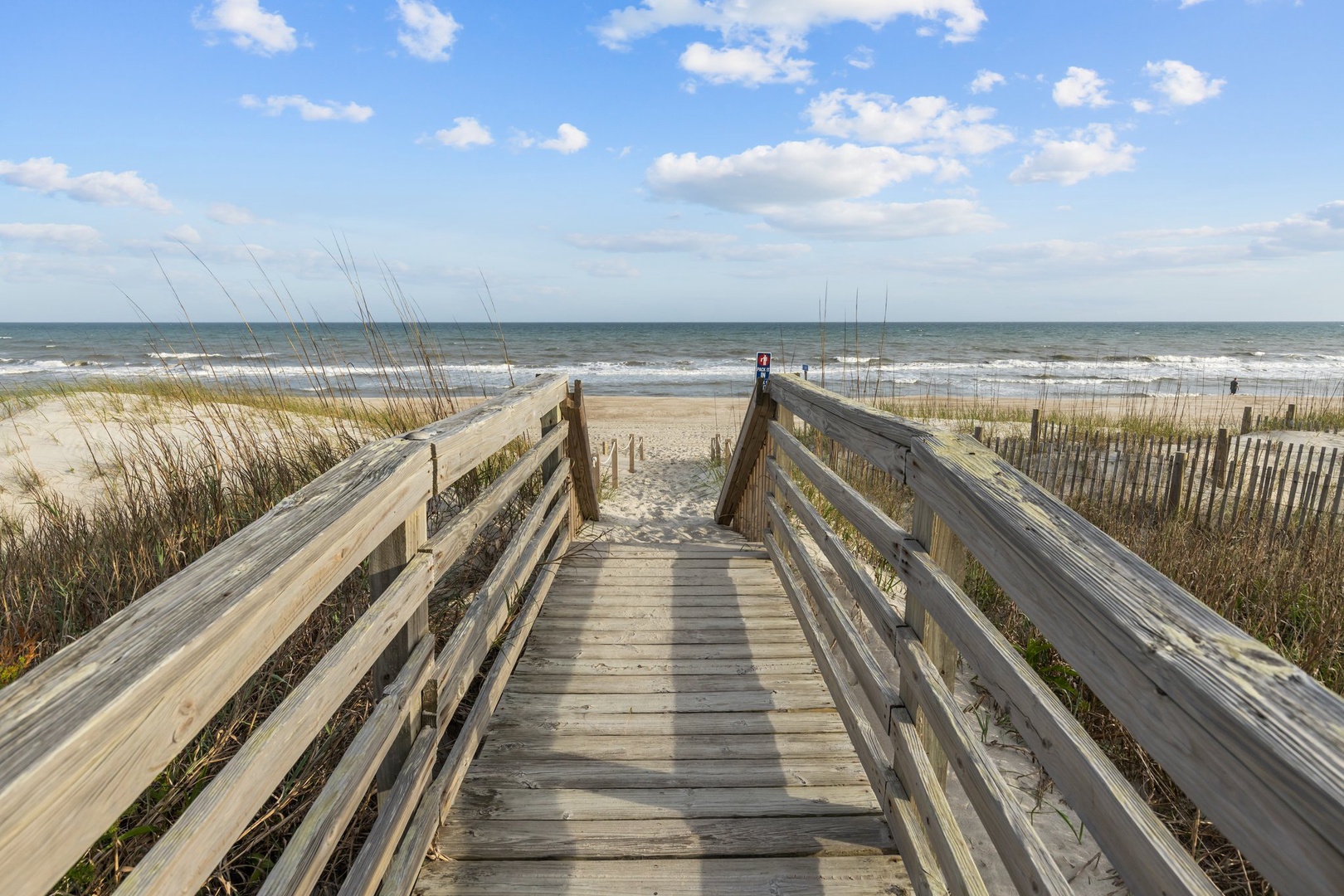 Wooden boardwalk leads through dunes to pristine sandy beach with rolling waves and endless ocean views.