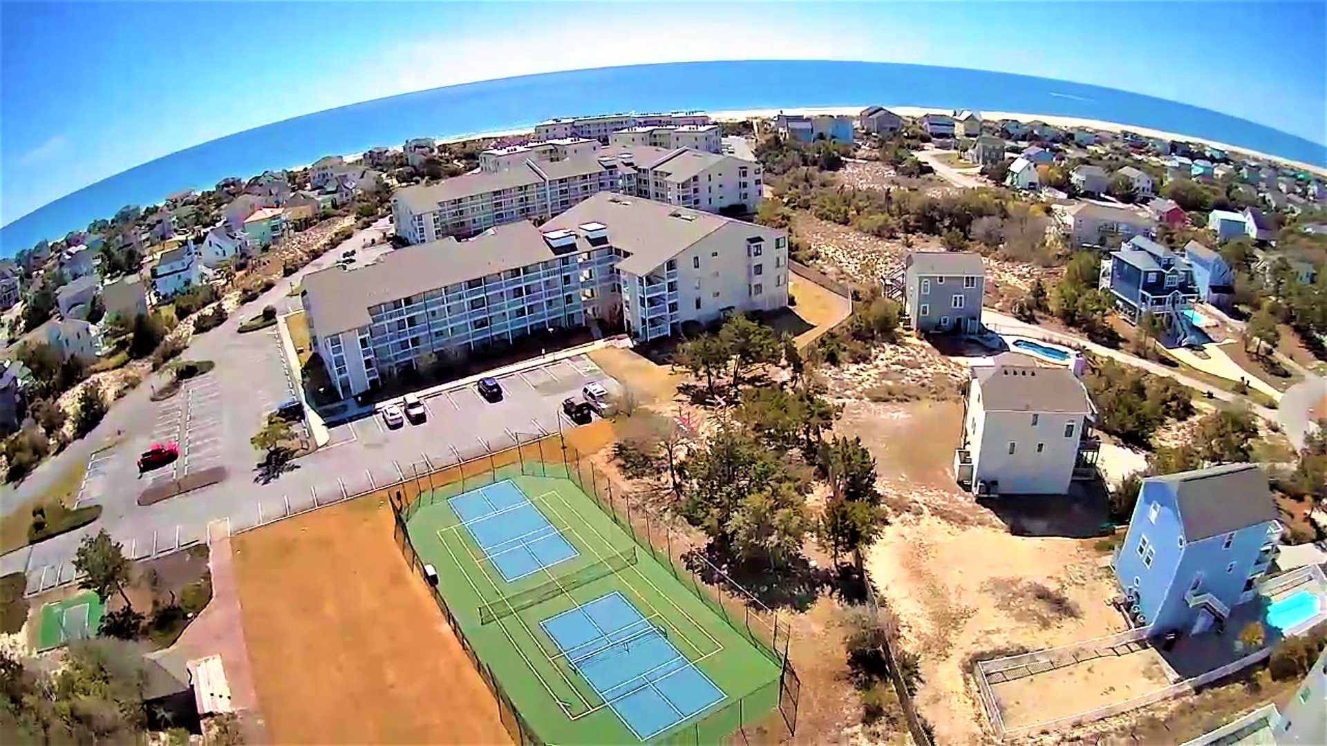 Aerial view of coastal vacation resort featuring tennis courts, multi-story accommodations, and beachside residential neighborhood.
