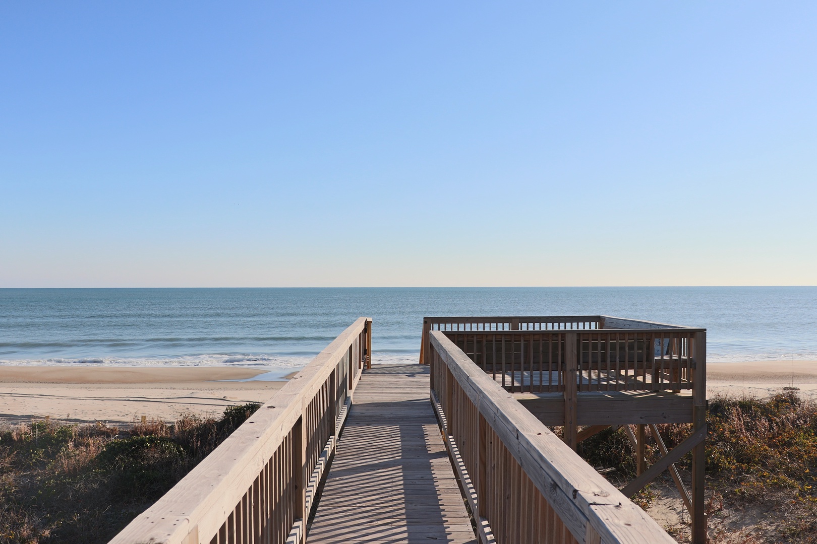 Wooden boardwalk leads to an elevated viewing platform overlooking pristine sandy beach and endless ocean horizon under clear blue skies.