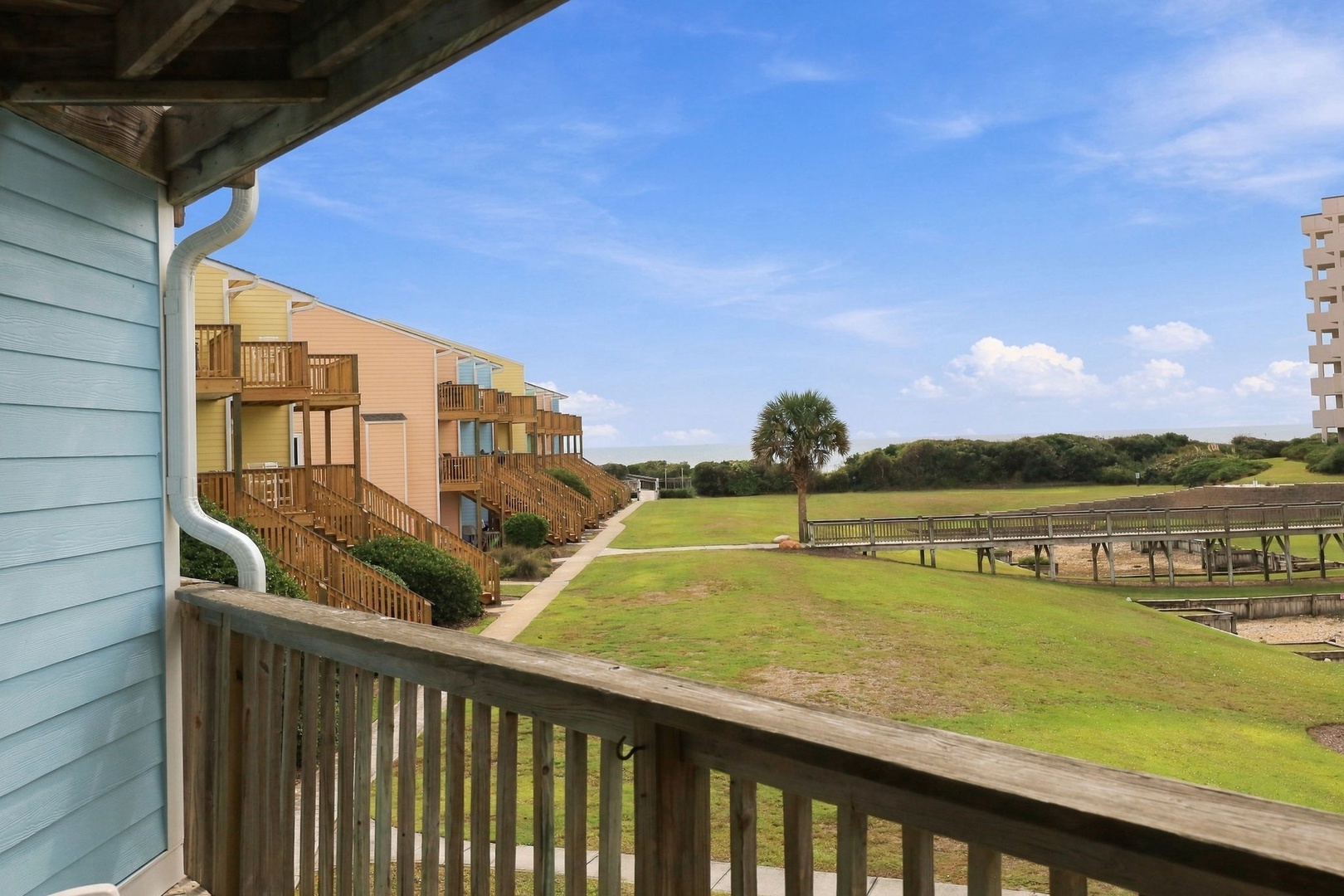 The property features exterior walkways connecting colorful beach-style units surrounded by lush coastal greenery and open grassy areas leading toward natural dunes.
