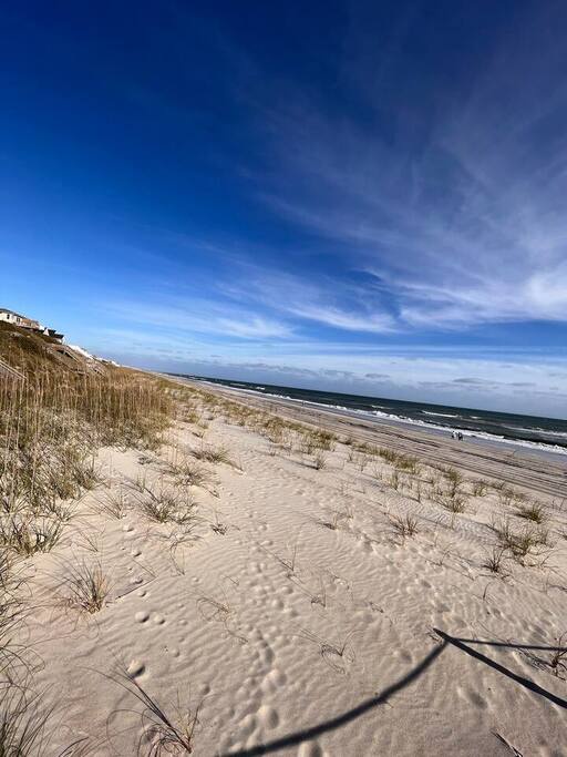 Pristine sandy coastline stretches beneath dramatic blue skies, with gentle dunes and beachside properties creating a perfect coastal setting.