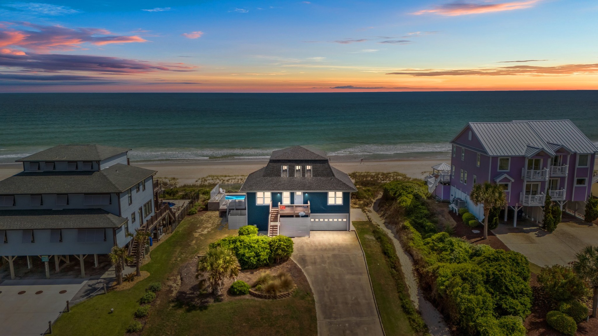 Elevated beachfront homes line the pristine coastline under a breathtaking sunset sky with vibrant coral and purple hues stretching across the horizon.