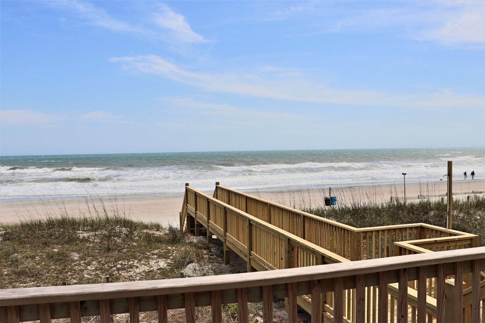Wooden boardwalk leads to pristine sandy beach with rolling waves under expansive blue skies.