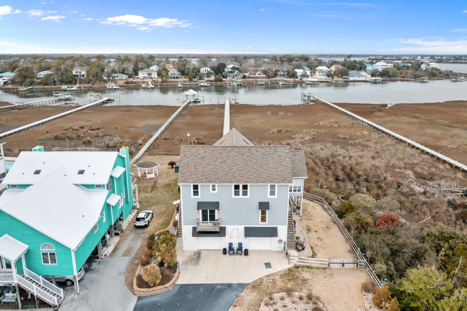 Aerial view of coastal vacation rental with waterfront access via private pier. Property features multiple levels and sits among marsh and residential neighborhood.