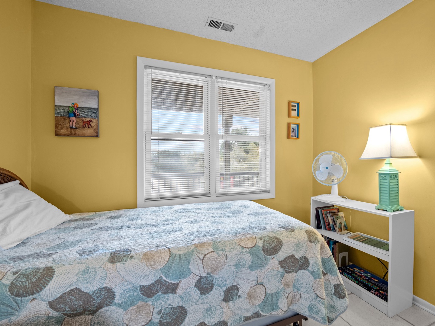 Relax in your cozy bedroom with sunny yellow walls, coastal-themed bedding, and thoughtful touches like a reading lamp and fan for comfort.