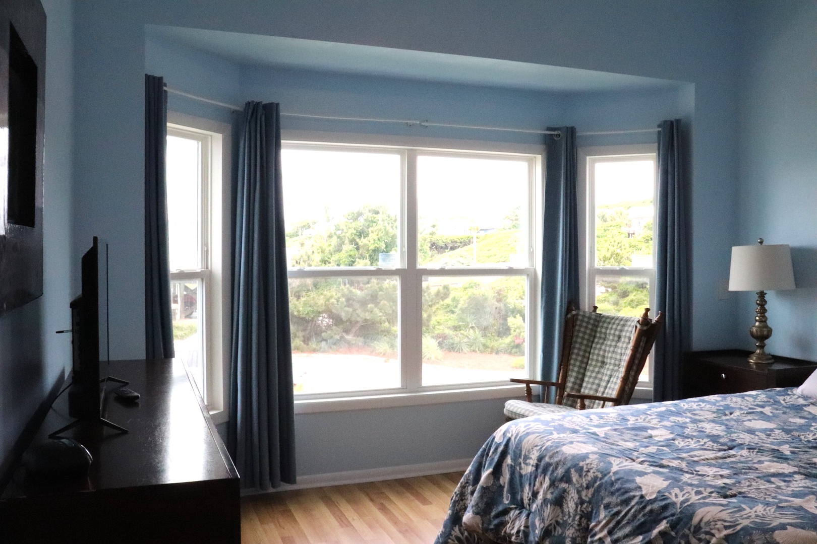 Natural light fills your serene bedroom, where peaceful blue walls and soft linens create a calming retreat. Wake up to lush greenery and open skies framed by large windows.