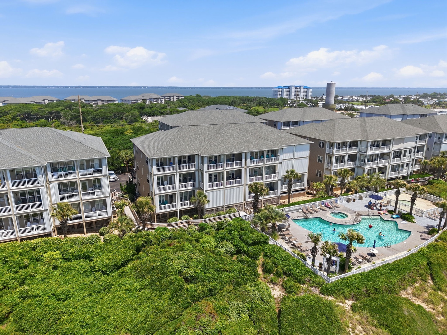 Aerial view of a coastal resort property featuring multi-story buildings with spacious balconies, a lagoon-style pool with hot tub, and lush tropical landscaping near the waterfront.