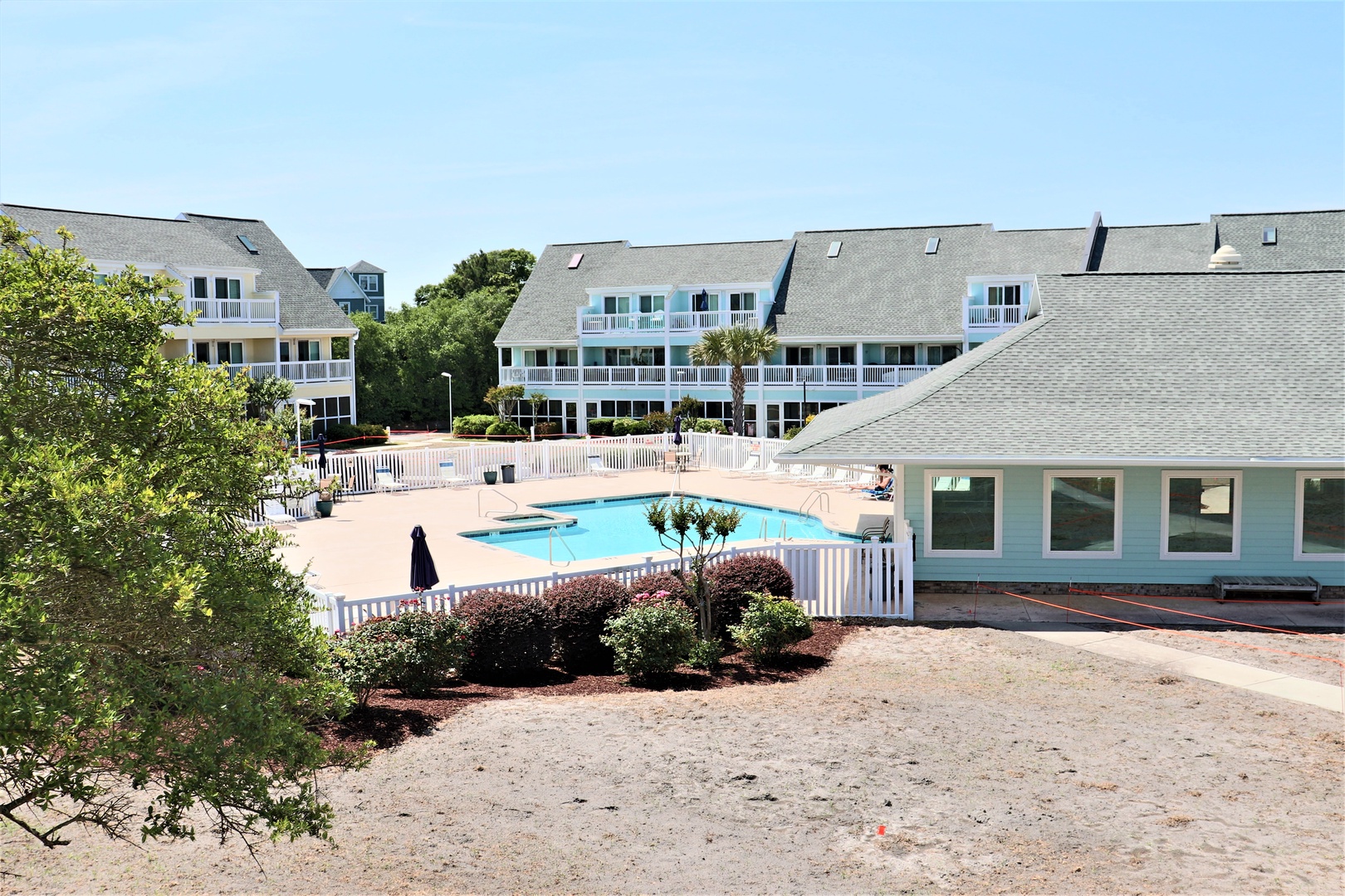 Coastal resort complex featuring multiple buildings, landscaped grounds, and a central pool area with white fencing.