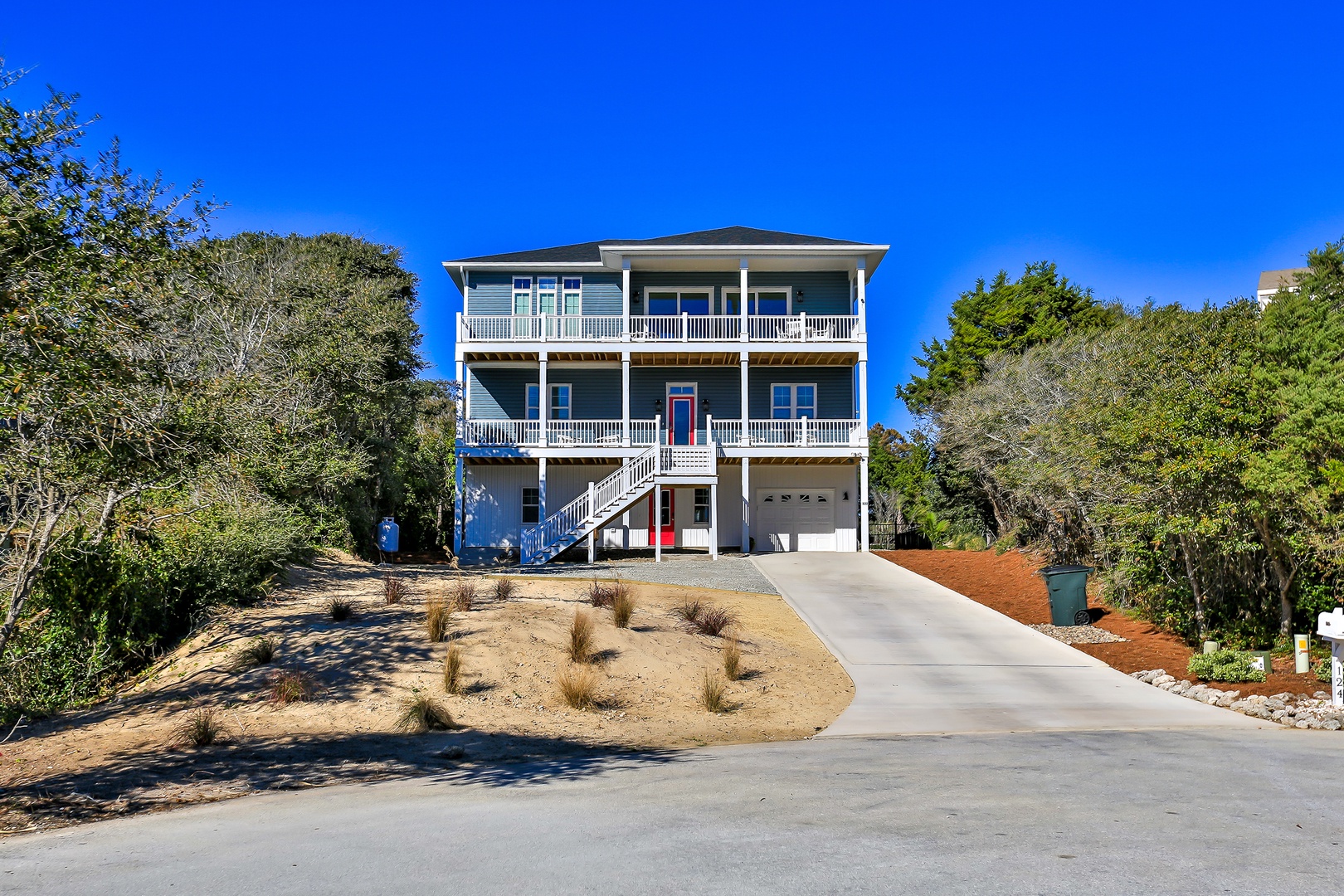 Charming three-story beach house with wraparound porches nestled among mature trees, featuring garage parking and coastal landscaping.