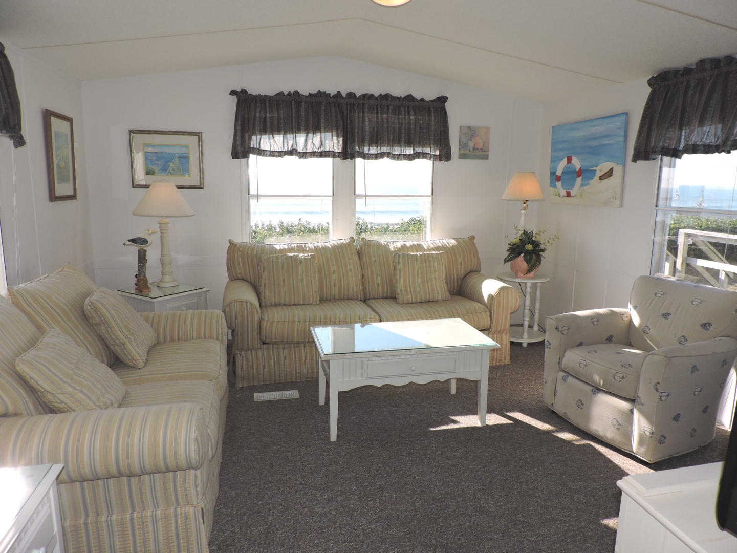 Settle into your bright living room where coastal charm meets comfort—striped sofas, nautical art, and windows overlooking peaceful scenery create the perfect retreat.