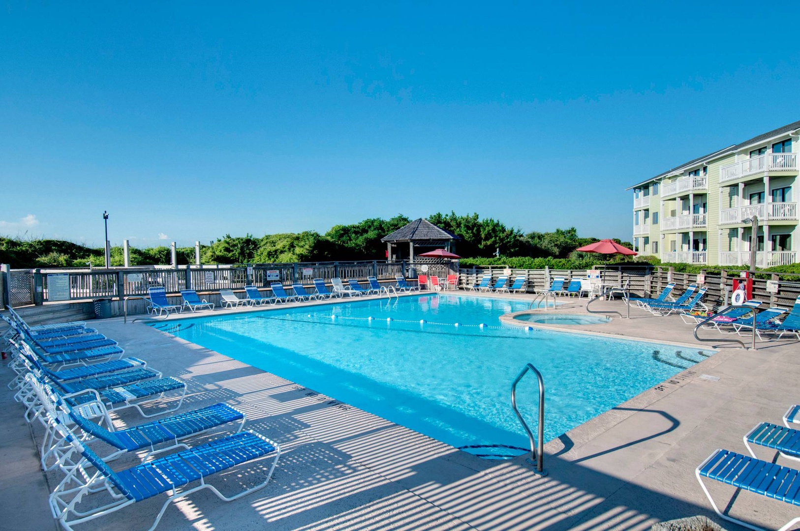 Large outdoor swimming pool surrounded by comfortable loungers under clear coastal skies, with oceanfront buildings nearby.