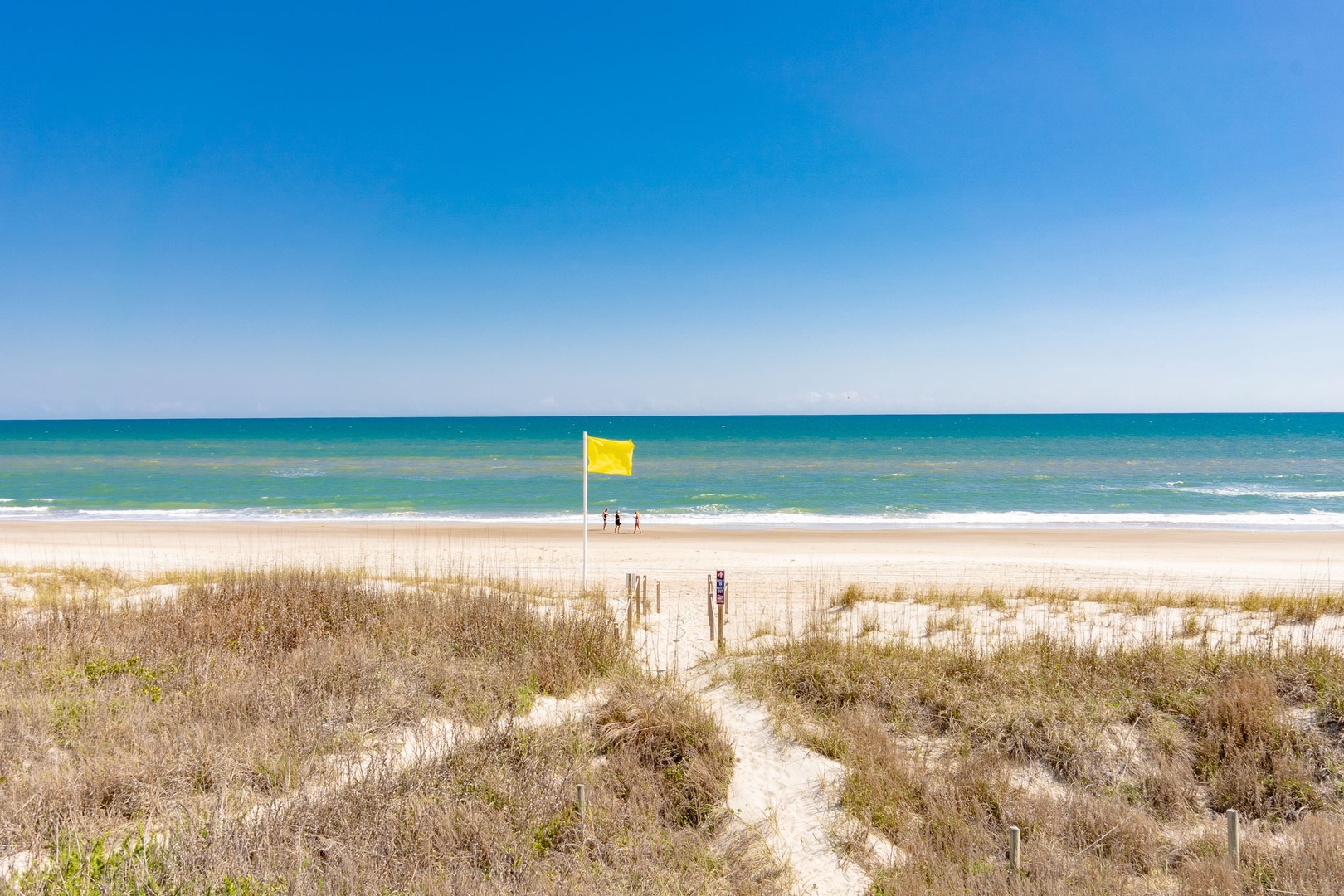 Pristine beach with crystal-clear waters and golden sand, featuring a yellow safety flag and peaceful dune pathway.
