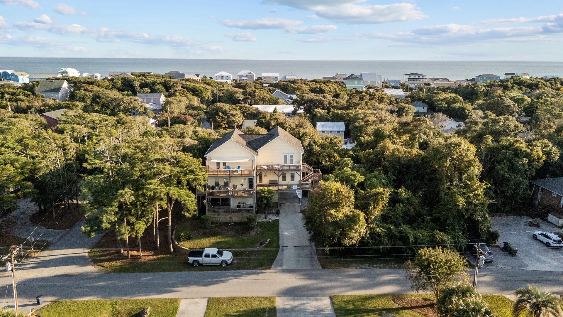 Aerial view of a coastal vacation home nestled among lush trees, with the ocean and nearby beach houses visible in the distance.