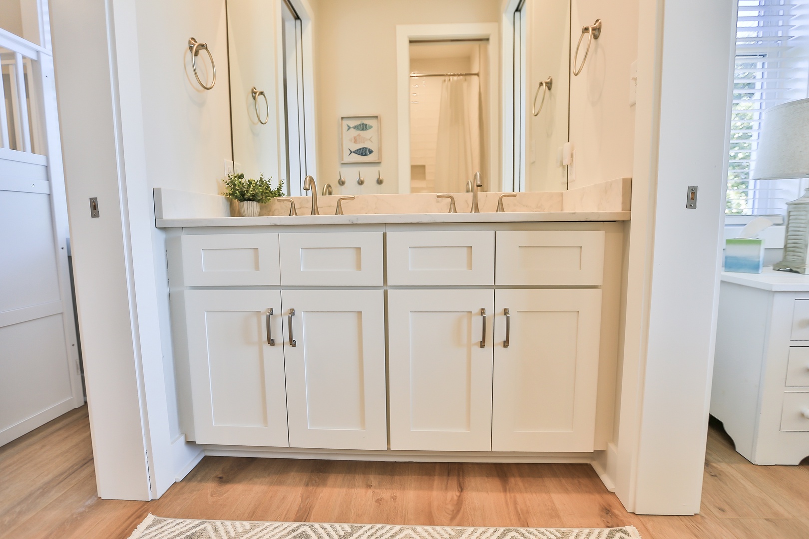 Unwind in this bright, spacious bathroom featuring dual vanities, elegant white cabinetry, and warm wood floors for your comfort.