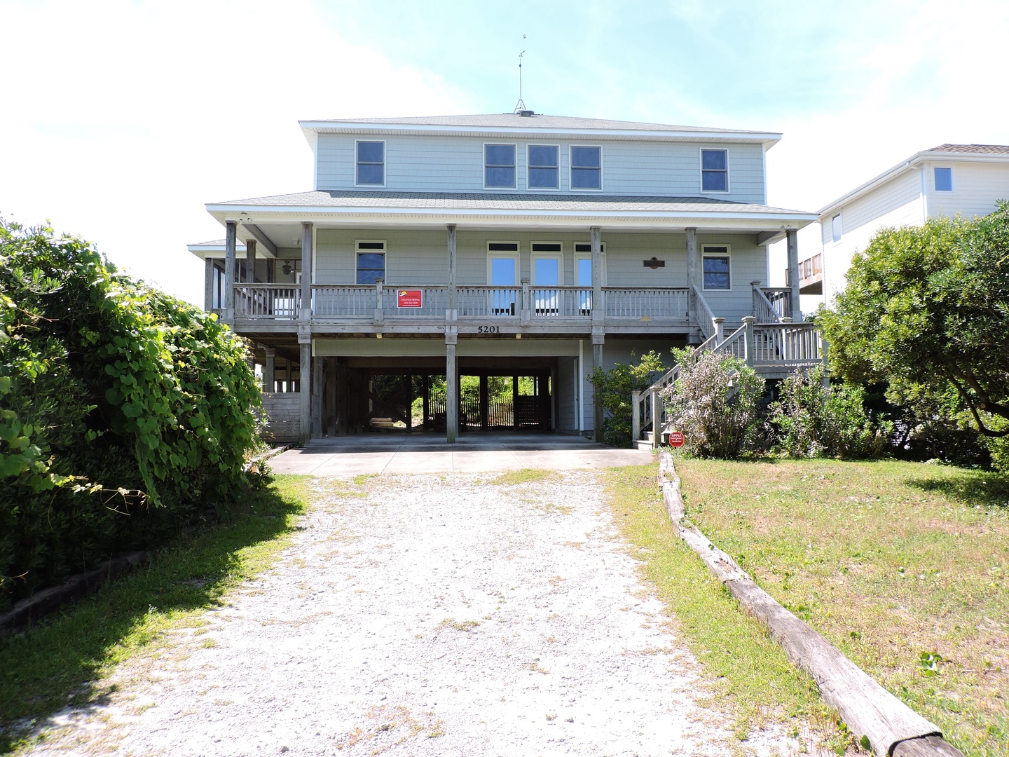 Two-story coastal vacation home featuring ground-level parking, spacious covered porch, and classic beach house architecture in quiet residential setting.