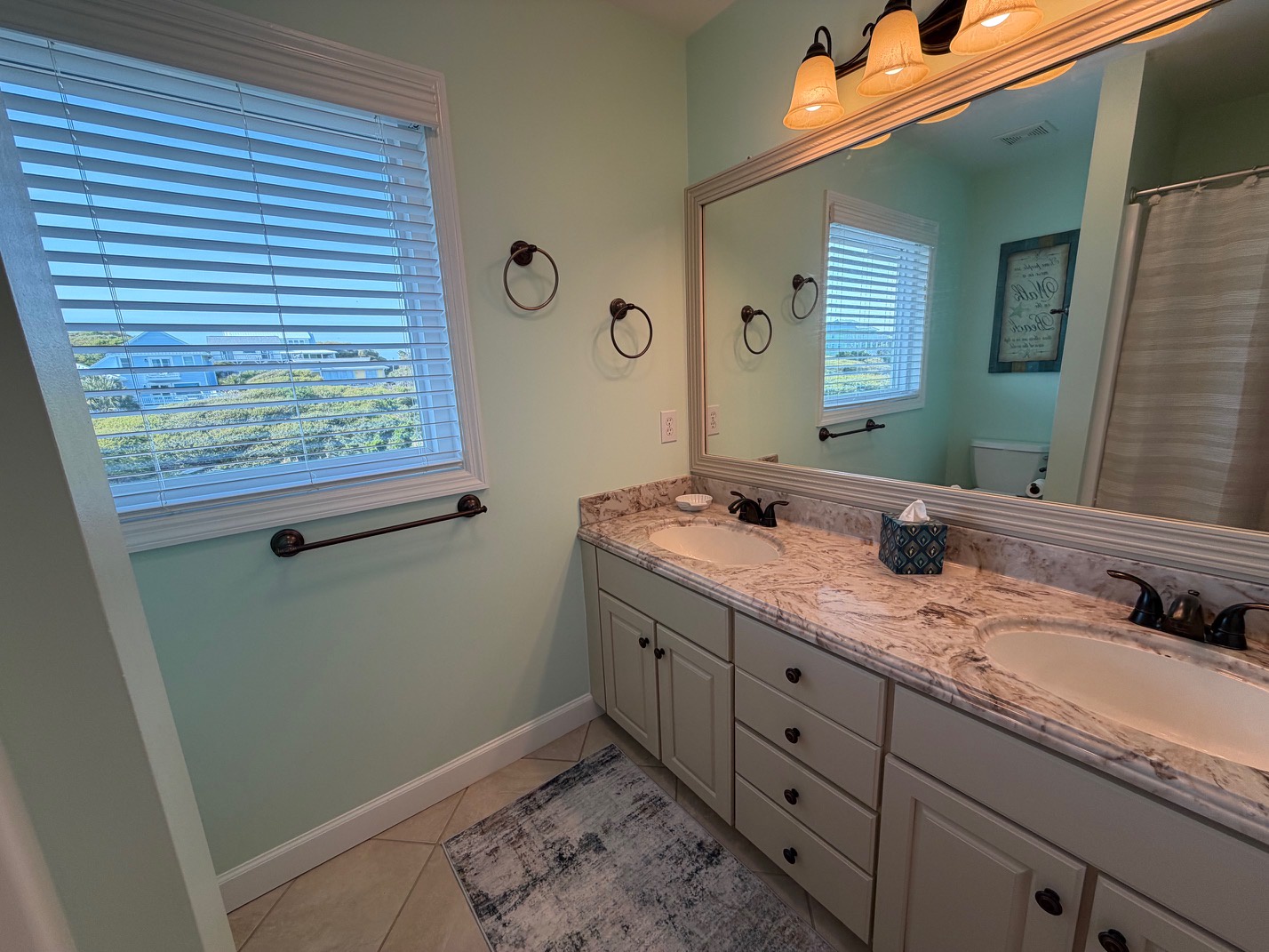 Refresh in your elegant dual-sink bathroom with marble countertops, warm lighting, and peaceful garden views through blinds.