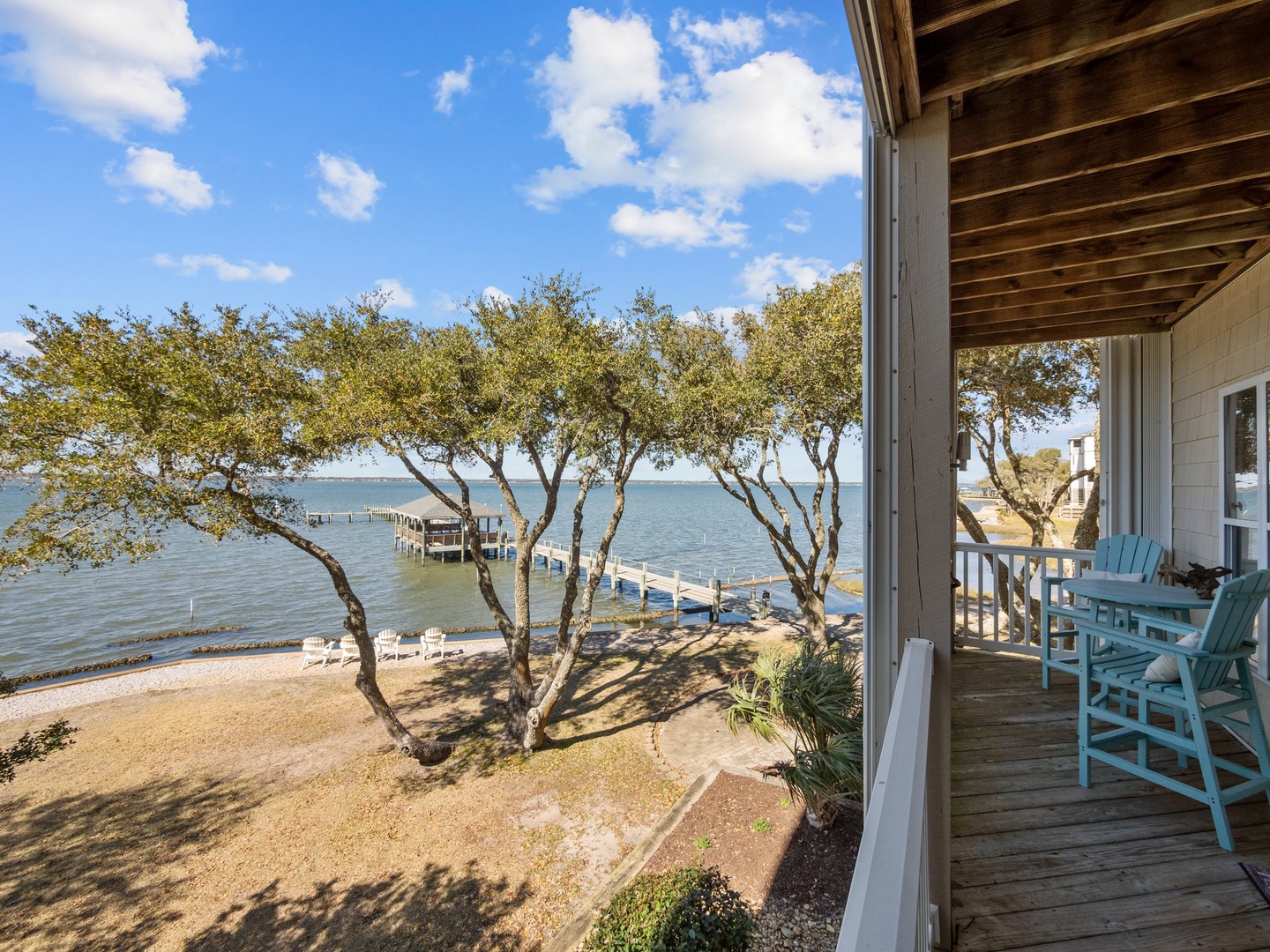 Waterfront balcony with turquoise chairs overlooks pristine bay waters, private dock, and sandy shoreline beneath mature trees.