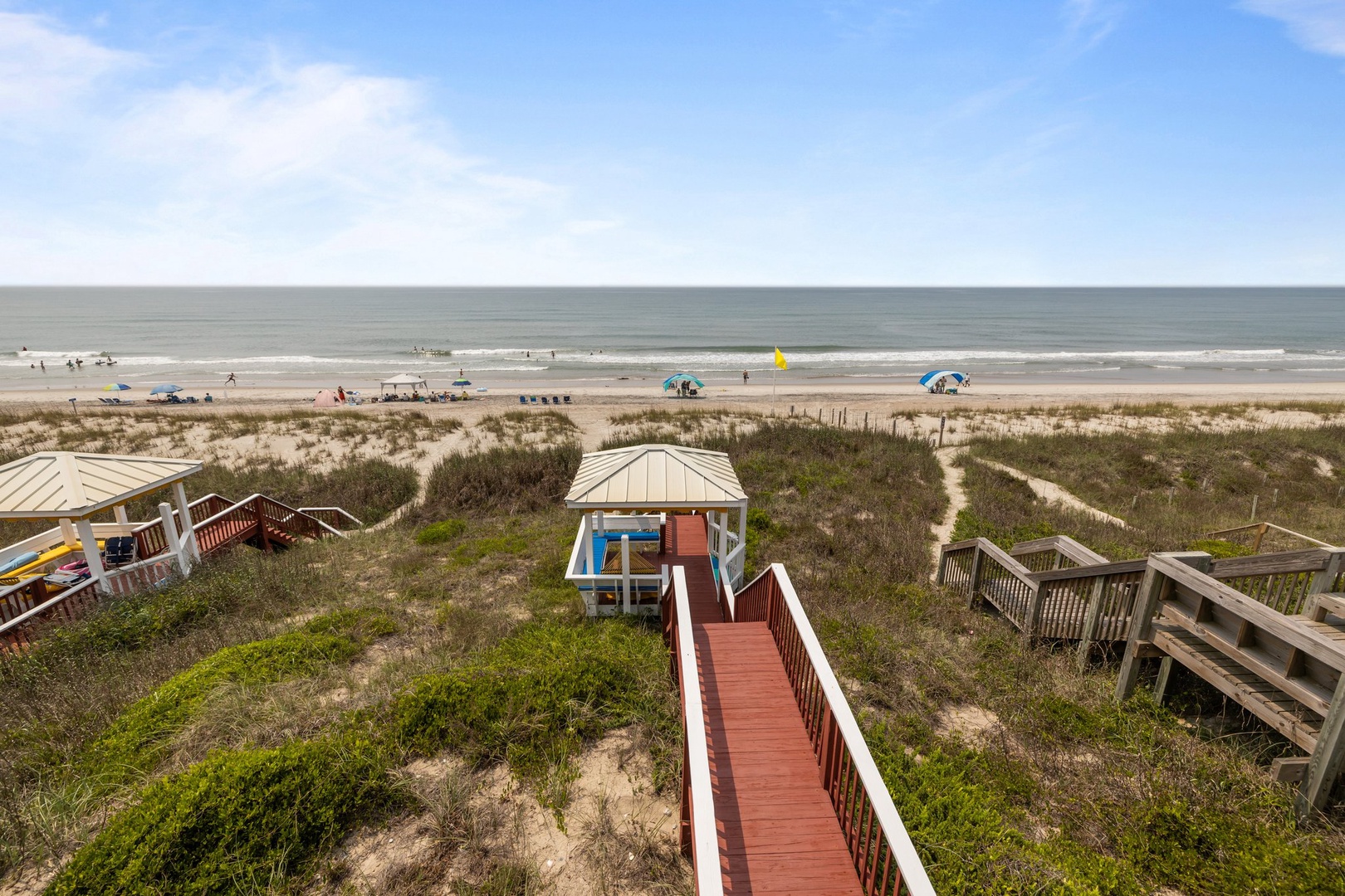 A wooden boardwalk leads through coastal dunes to a beautiful sandy beach with ocean views and beachgoers enjoying the shore.