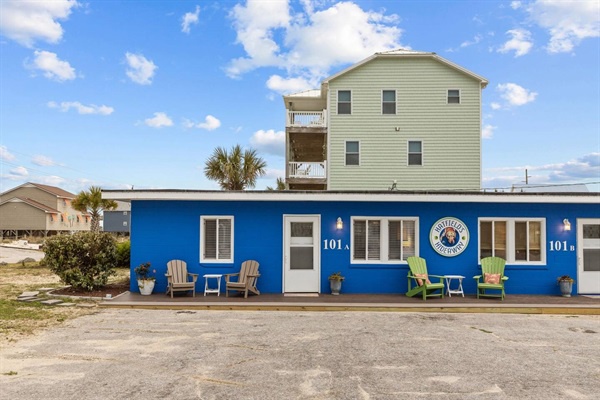 Bright blue beachside motel with cheerful Adirondack chairs and coastal charm welcoming guests to their seaside retreat.
