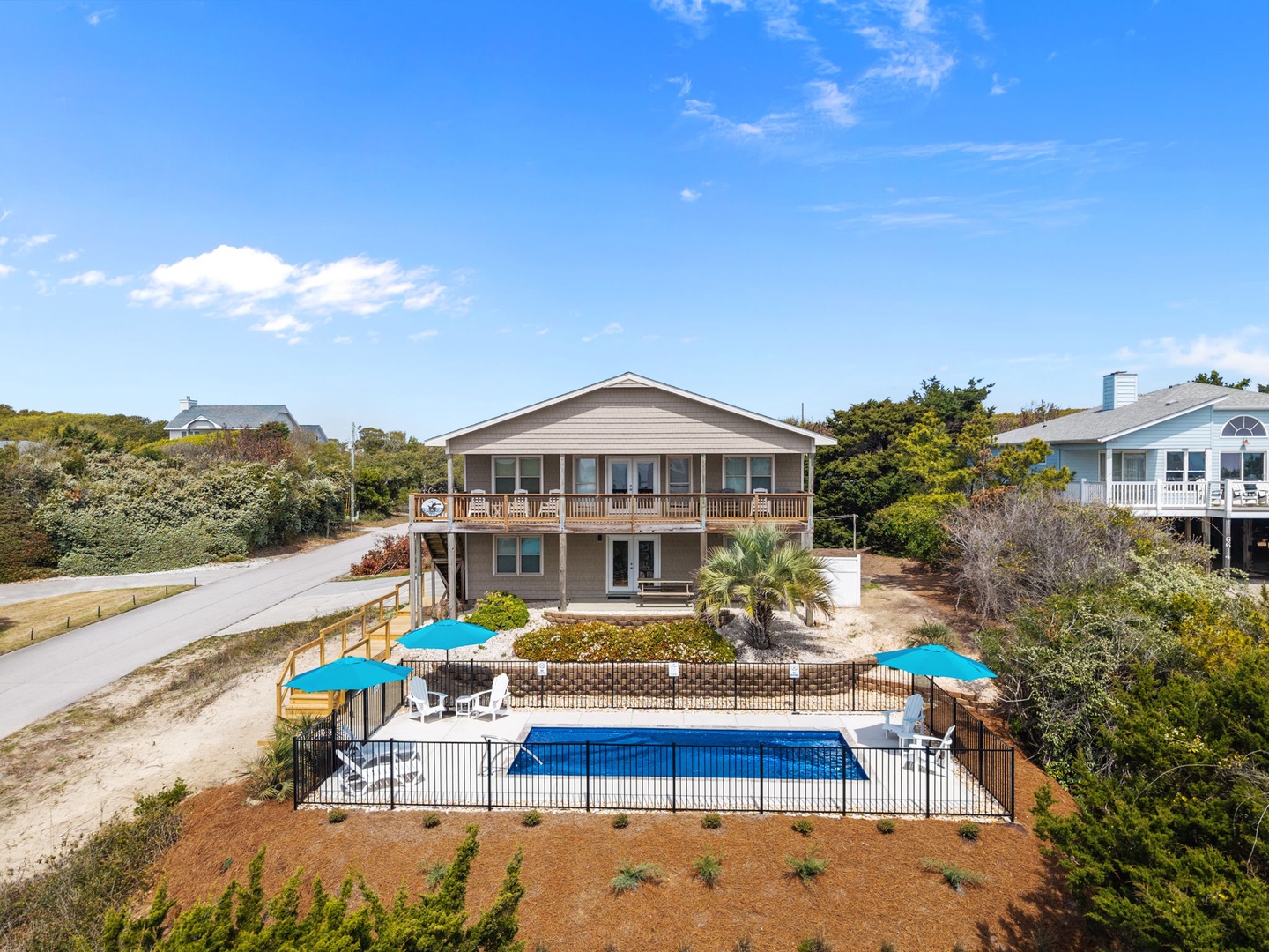 Beachside vacation home features private pool with safety fence, spacious deck with blue umbrellas, and lush coastal landscaping.
