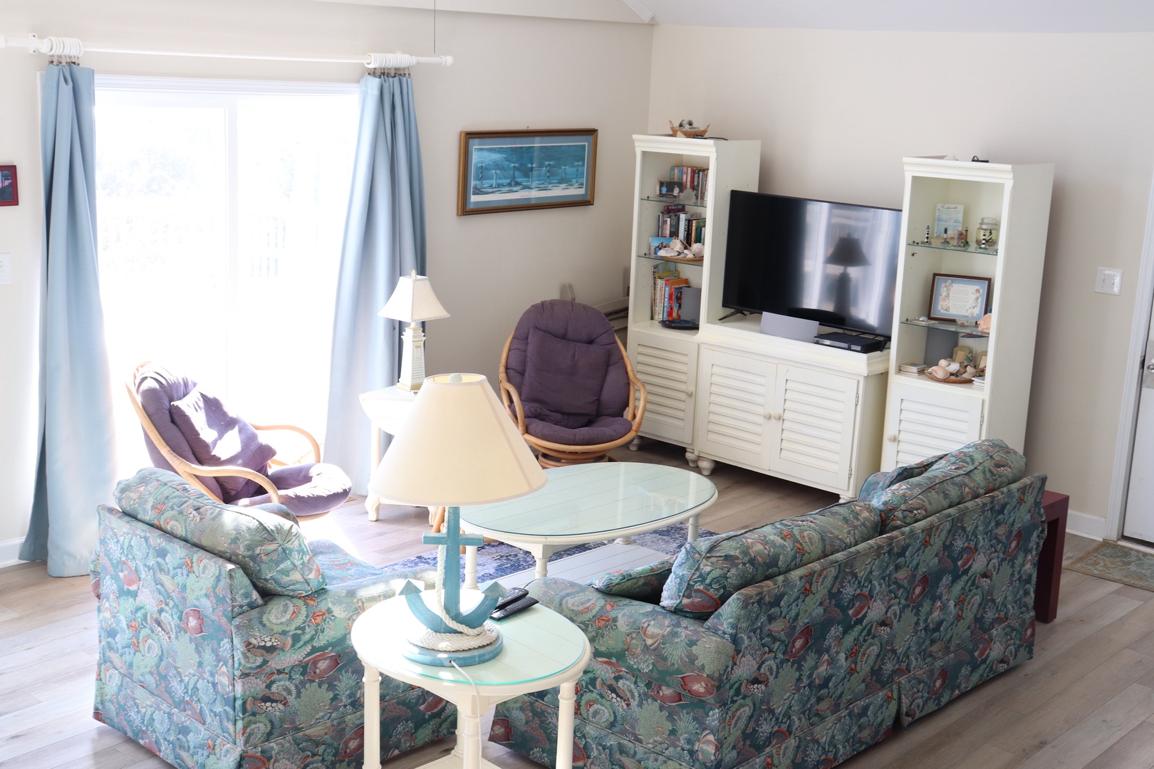 Unwind in your spacious living room with cozy floral seating, entertainment center, and bright natural light streaming through coastal-blue curtains.