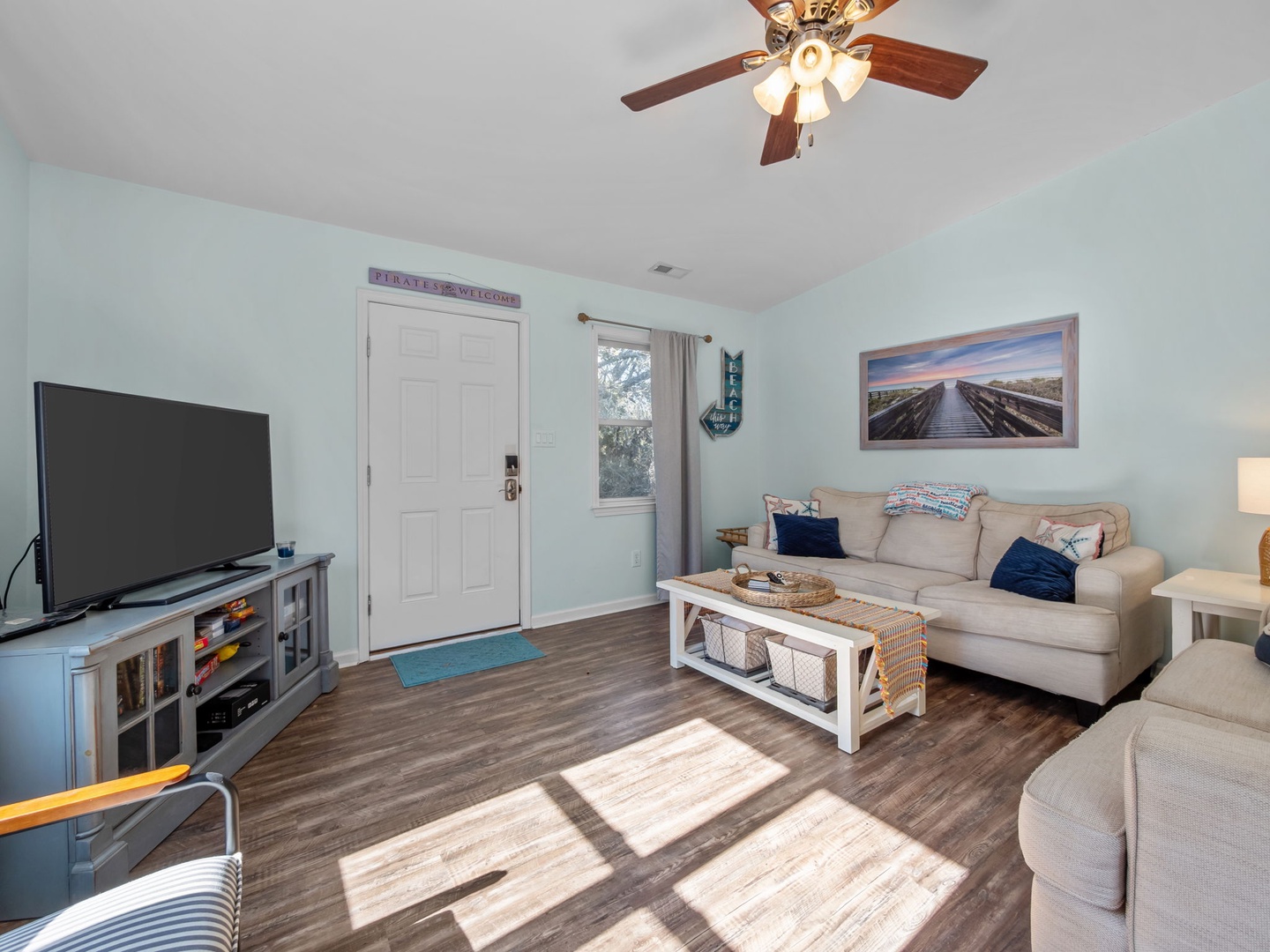 Unwind in your bright, coastal-themed living room with plush seating, beach artwork, and natural light streaming through windows.