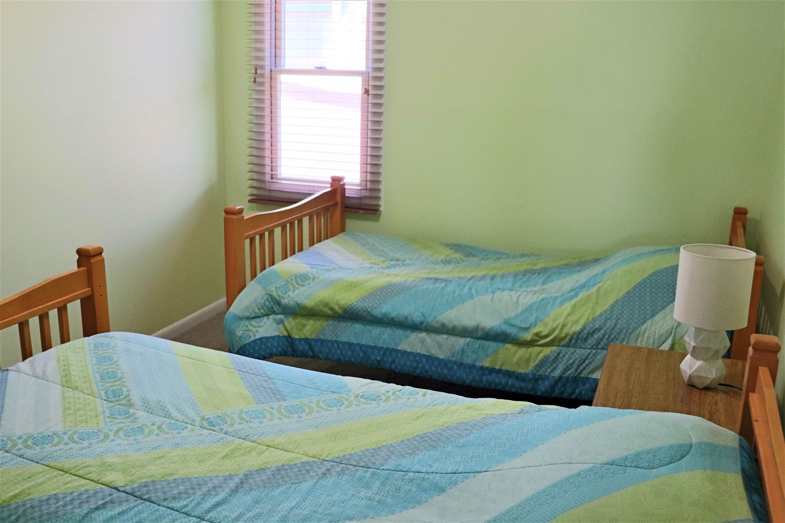 Rest peacefully in your bright bedroom with twin beds dressed in cheerful blue and green linens, natural light streaming through your window.
